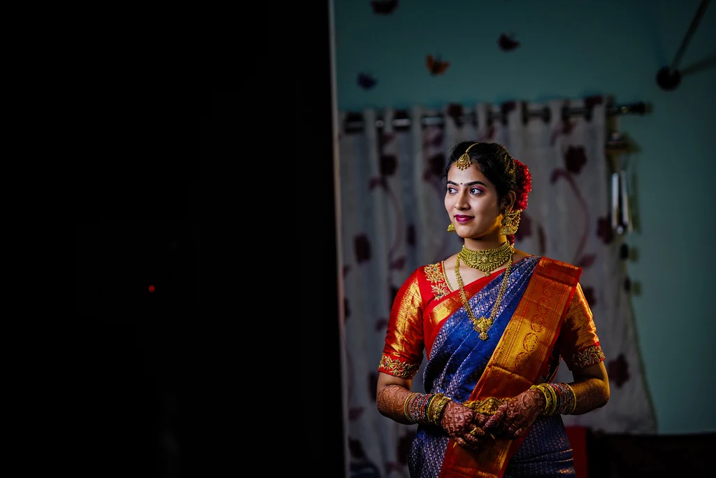 A bride in an orange and blue sari standing in a softly lit doorway